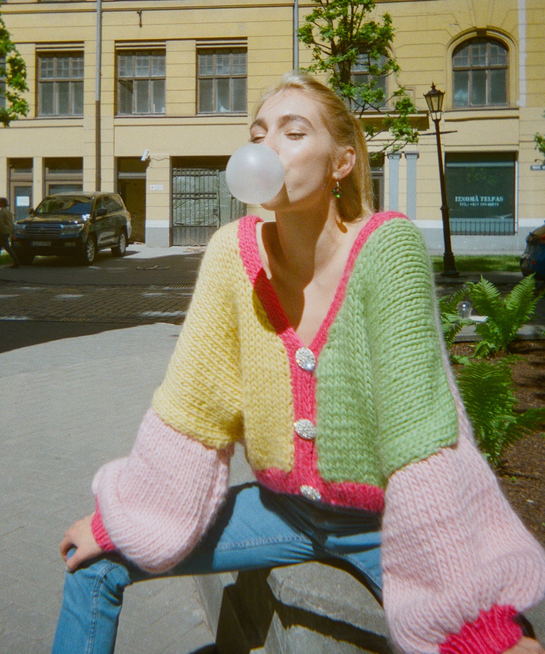 A woman is creating bubbles on a sidewalk wearing a colourful knitted cardigan.