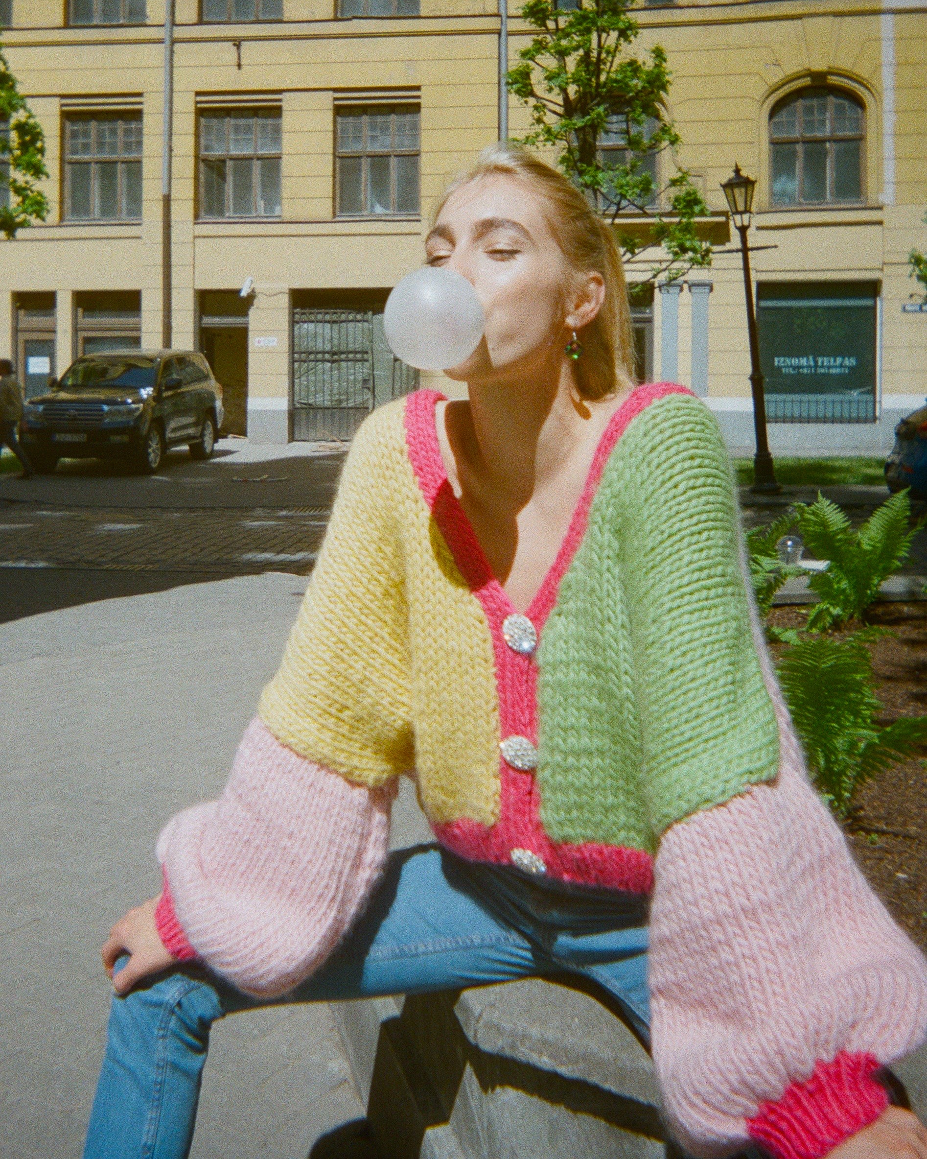 A woman is creating bubbles on a sidewalk wearing a colourful knitted cardigan.