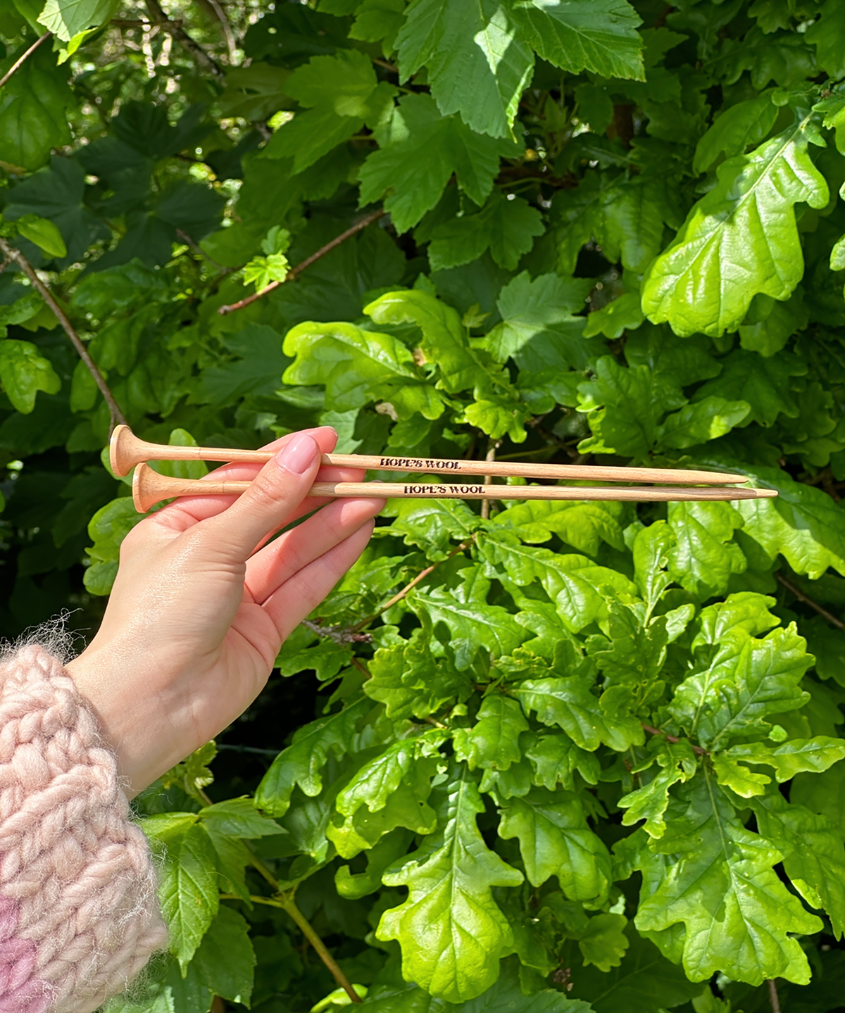 A person holding two wooden knitting needles in front of a bush, ready to start a new knitting project.