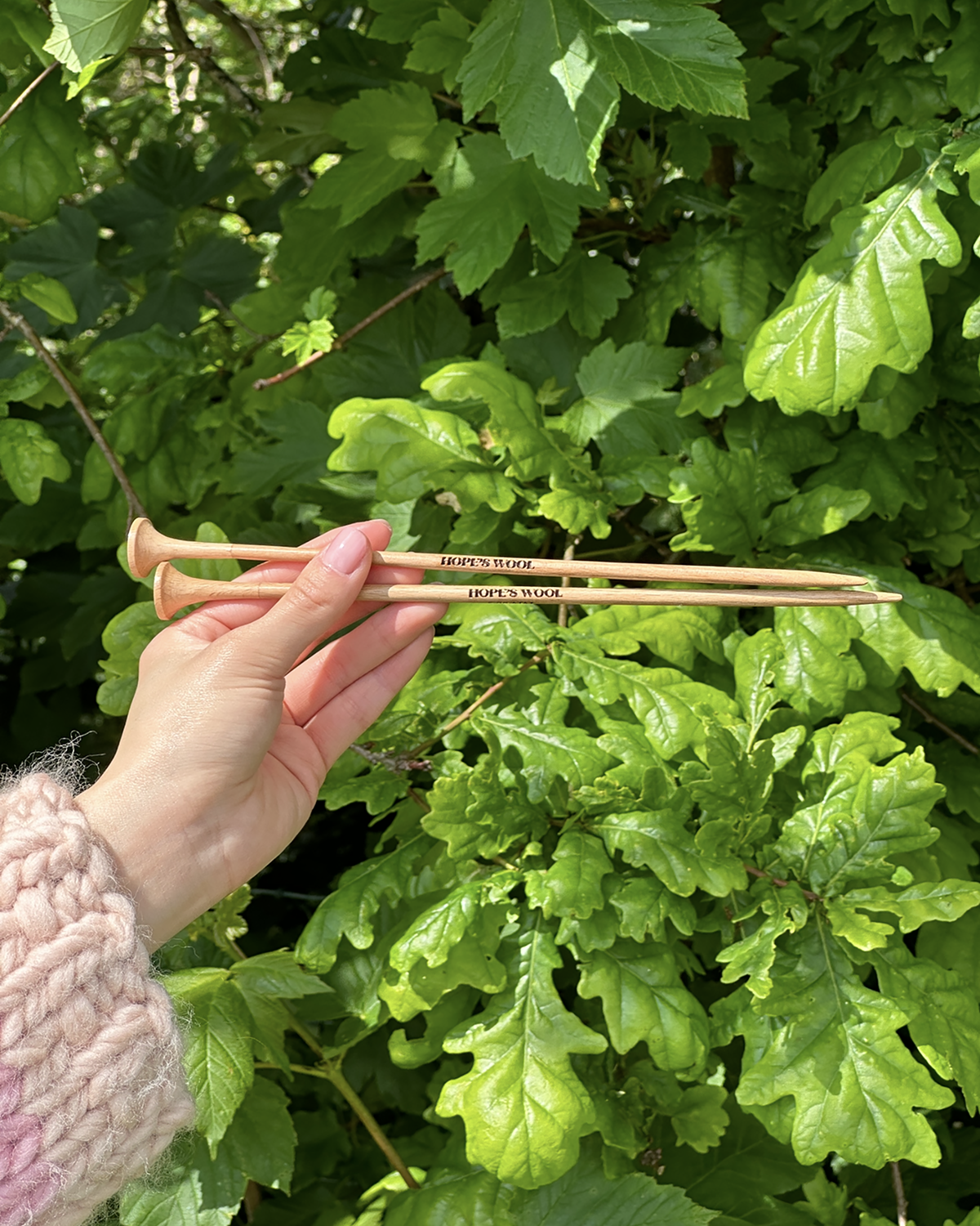A person holding two wooden knitting needles in front of a bush, ready to start a new knitting project.