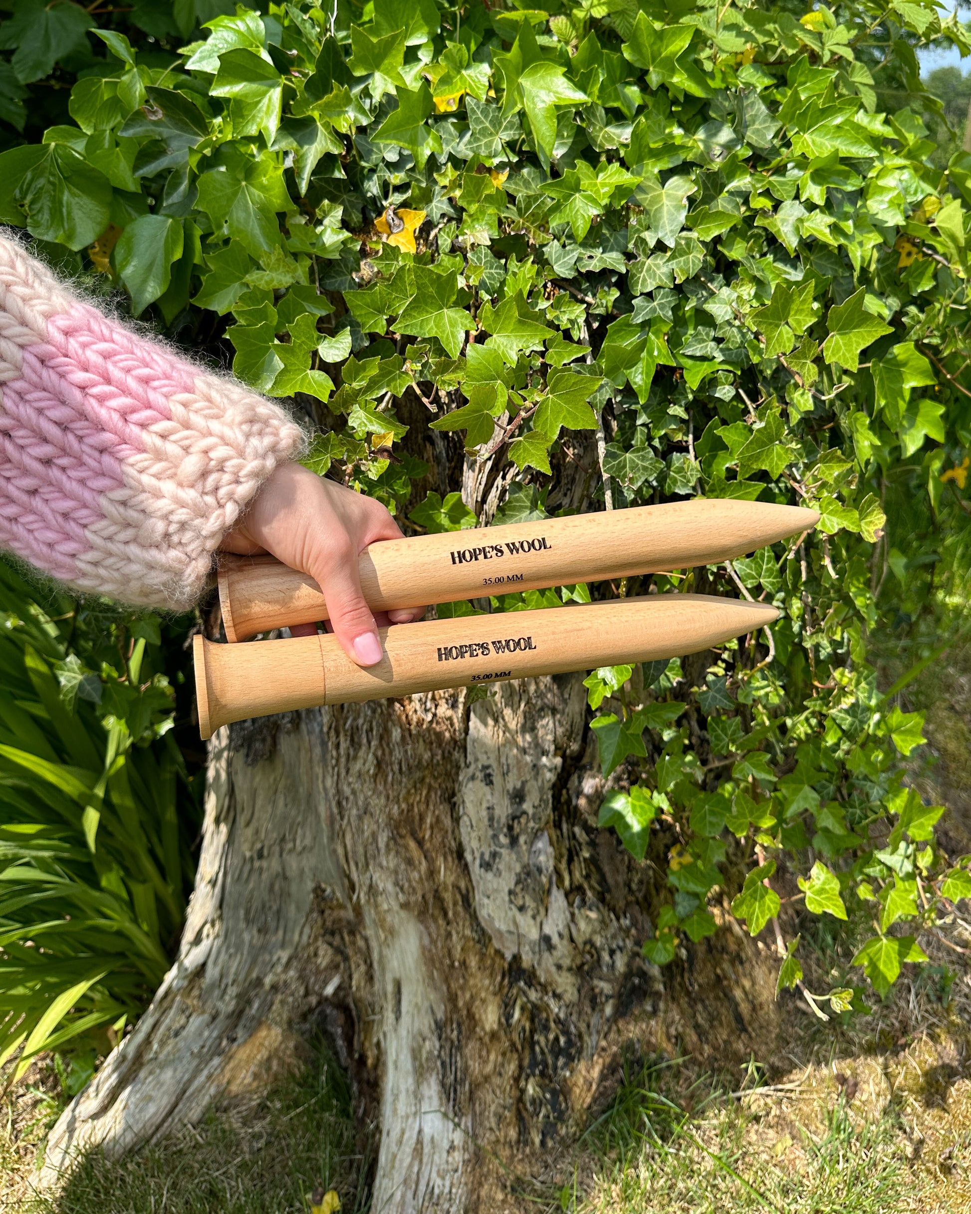 A person holding two wooden knitting needles in front of a bush, ready to start a new knitting project.