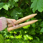 A person holding two wooden knitting needles in front of a bush, ready to start a new knitting project.