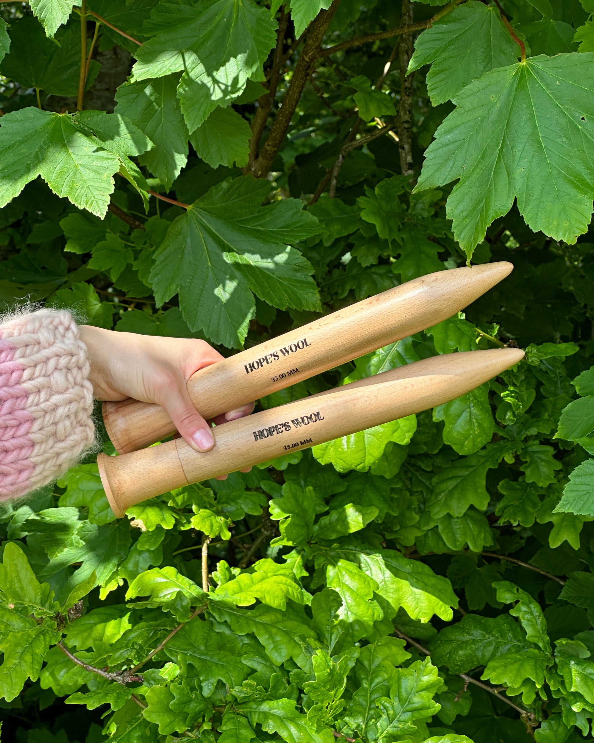 A person holding two wooden knitting needles in front of a bush, ready to start a new knitting project.