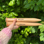 A person holding two wooden knitting needles in front of a bush, ready to start a new knitting project.