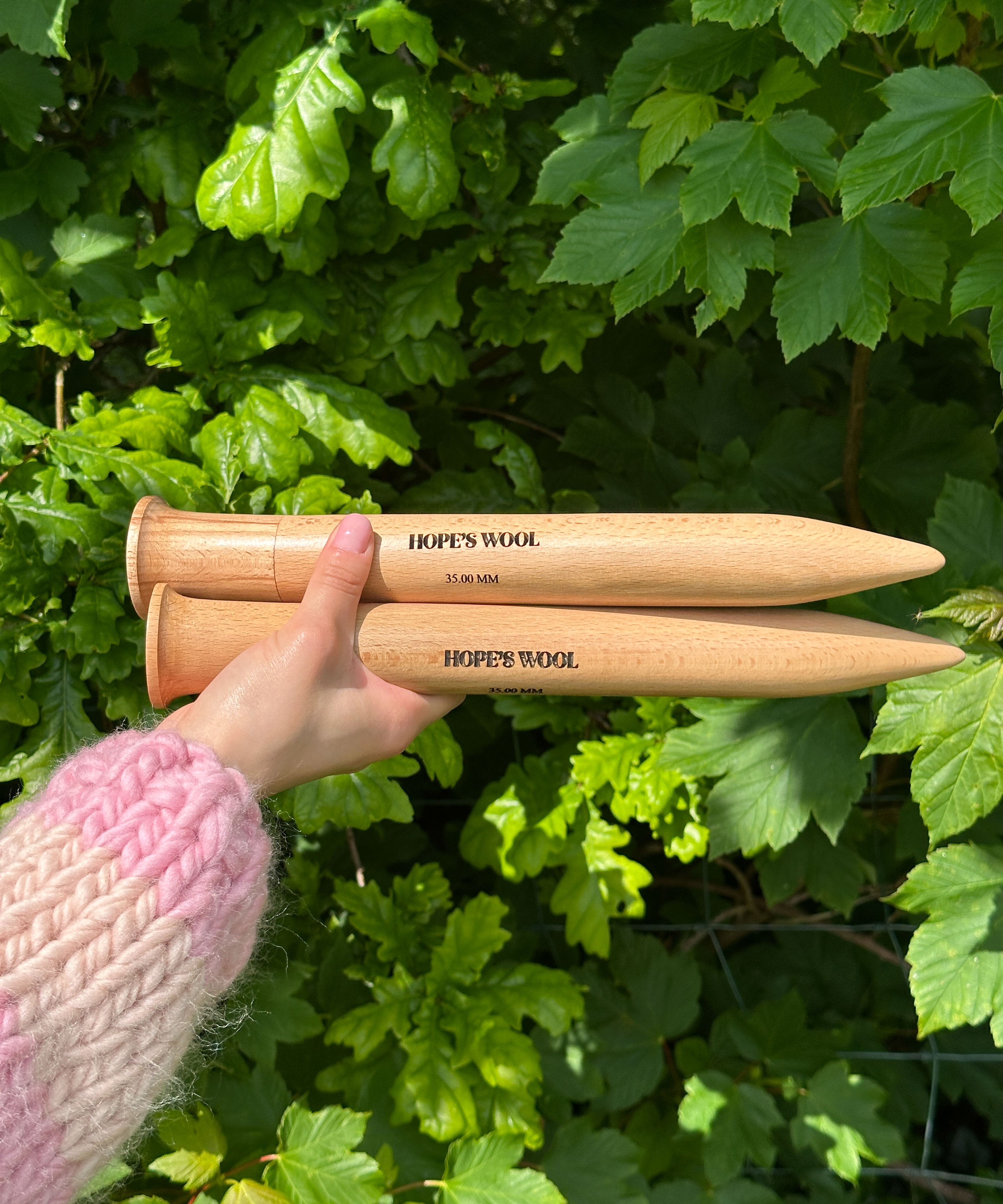 A person holding two wooden knitting needles in front of a bush, ready to start a new knitting project.