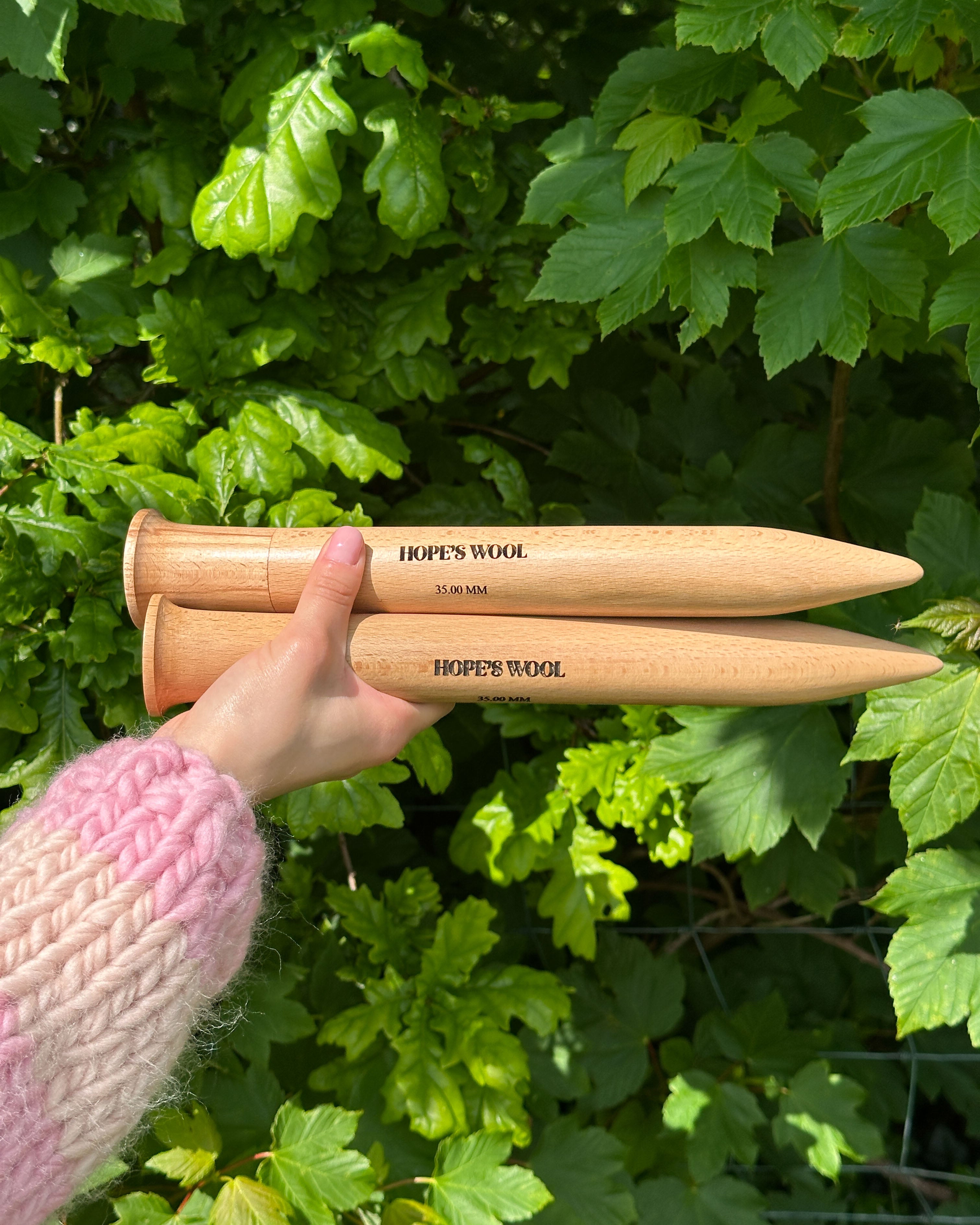 A person holding two wooden knitting needles in front of a bush, ready to start a new knitting project.