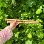 A person holding two wooden knitting needles in front of a bush, ready to start a new knitting project.