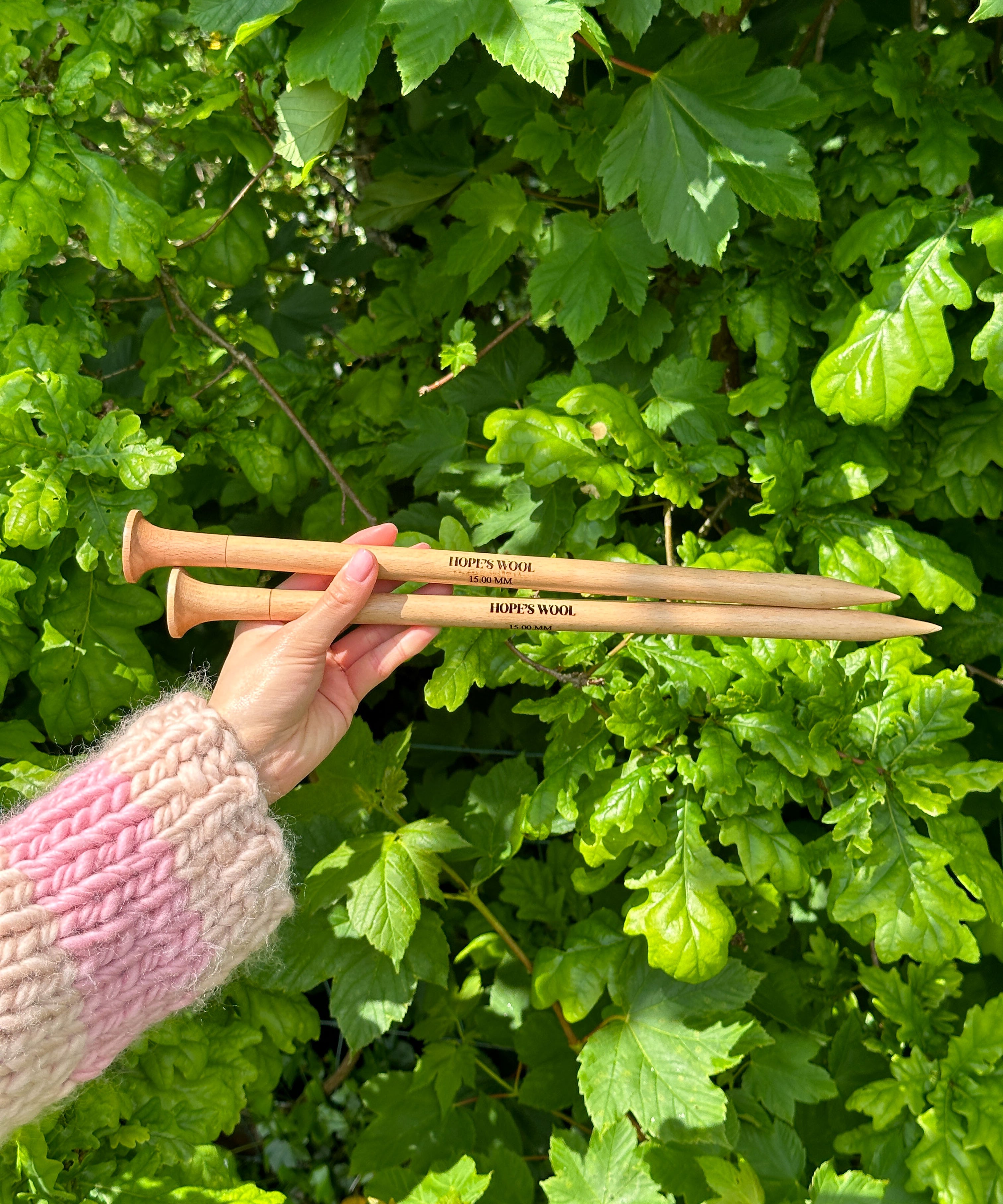 A person holding two wooden knitting needles in front of a bush, ready to start a new knitting project.