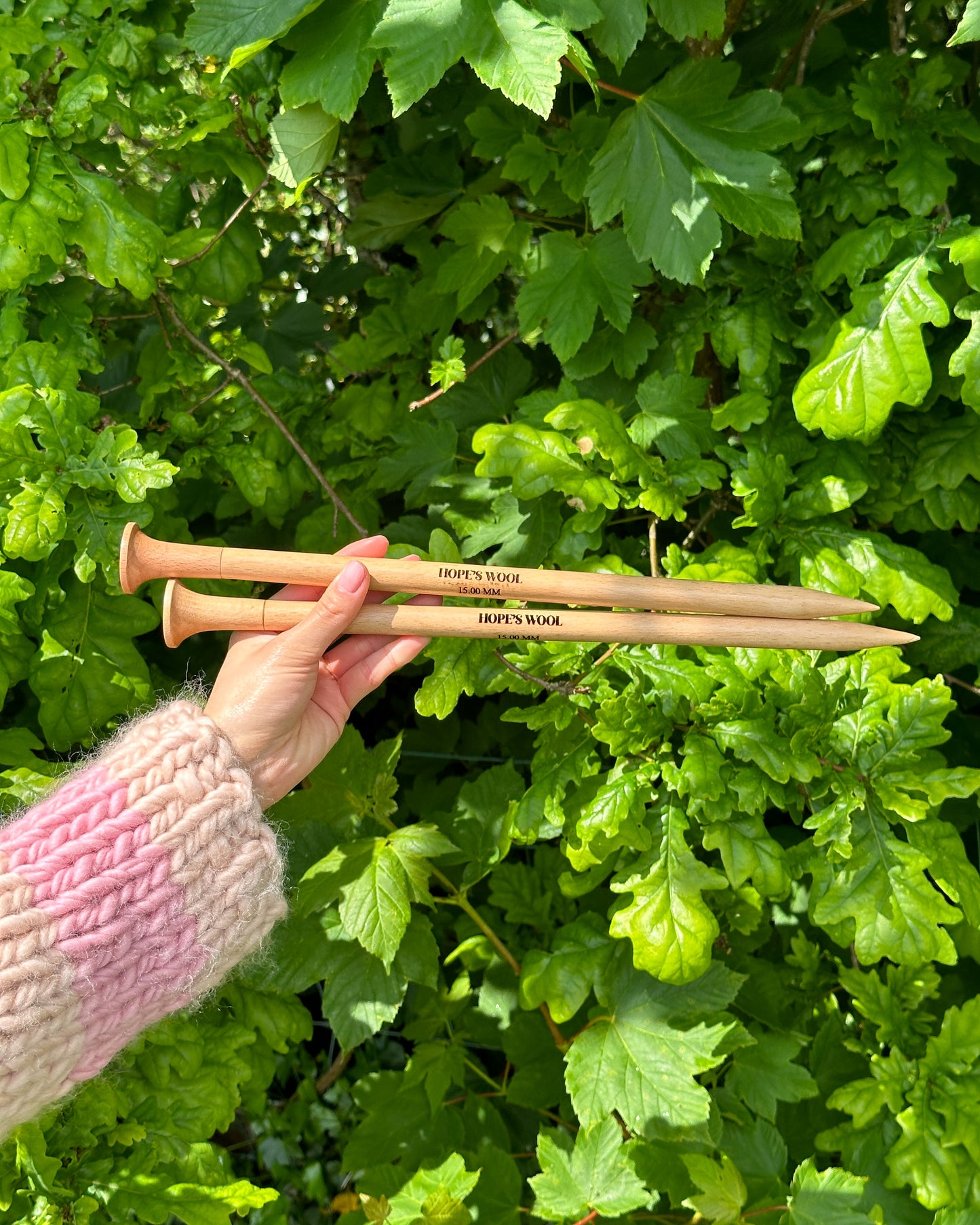 A person holding two wooden knitting needles in front of a bush, ready to start a new knitting project.