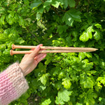 A person holding two wooden knitting needles in front of a bush, ready to start a new knitting project.