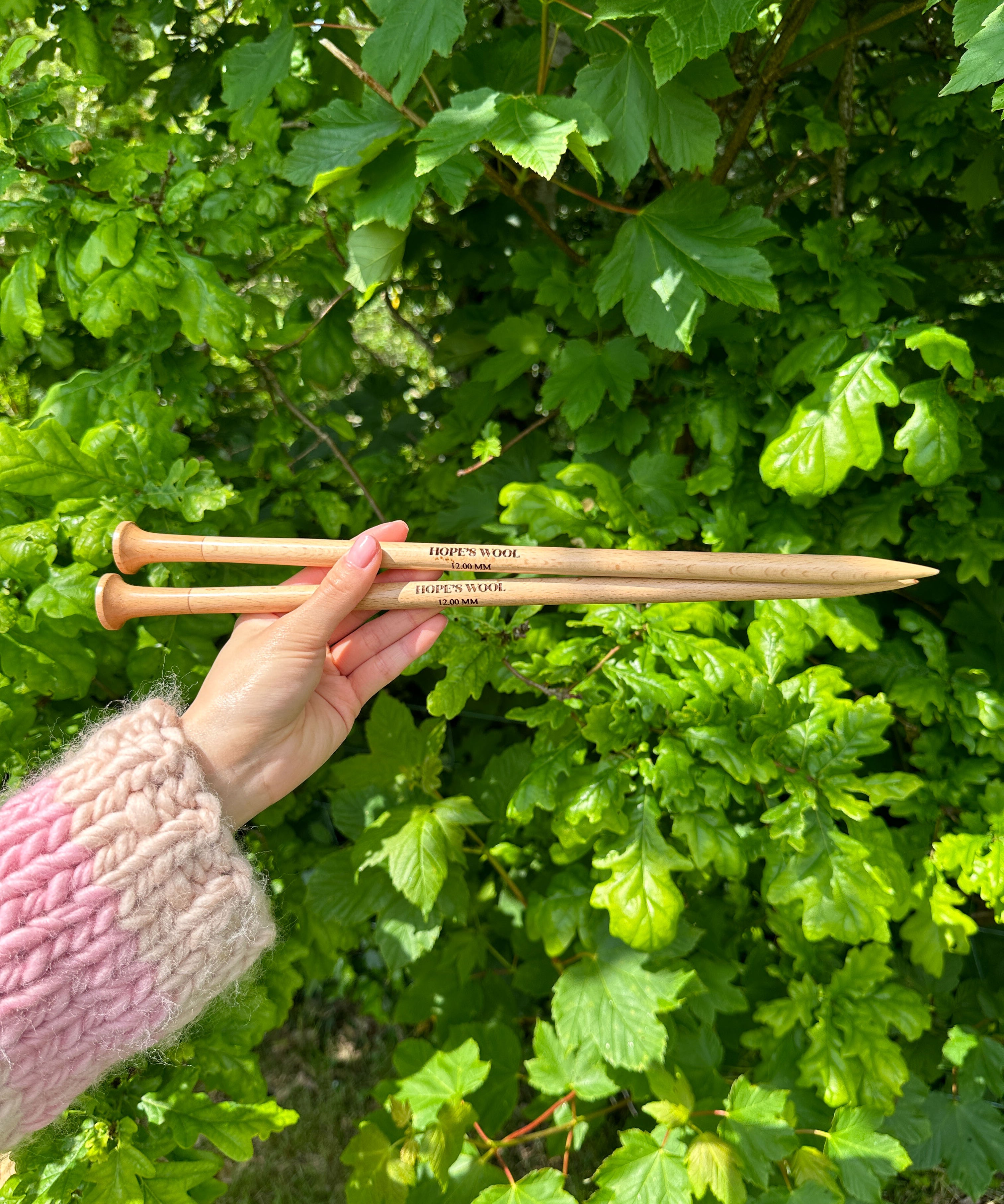 A person holding two wooden knitting needles in front of a bush, ready to start a new knitting project.