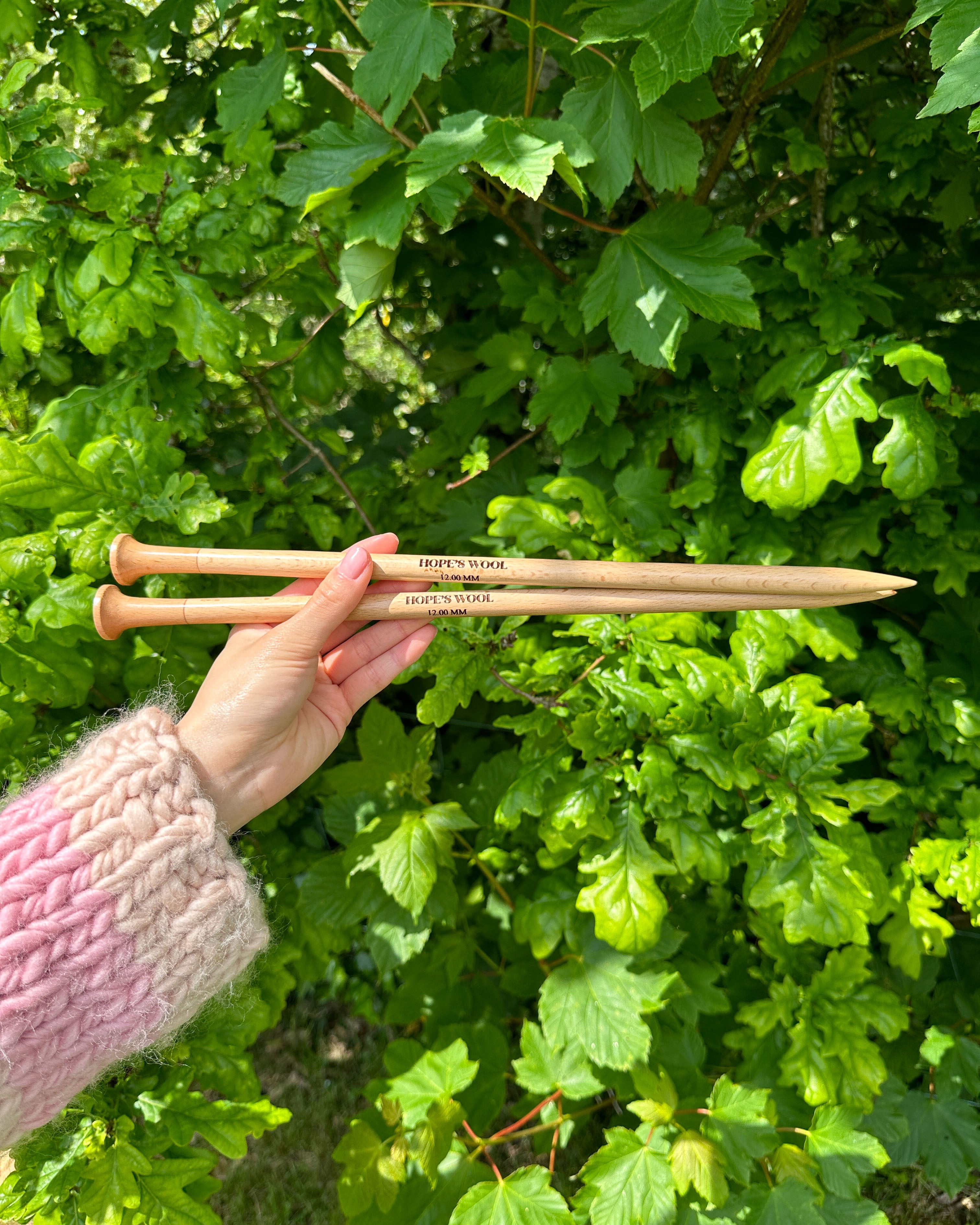 A person holding two wooden knitting needles in front of a bush, ready to start a new knitting project.