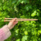 A person holding two wooden knitting needles in front of a bush, ready to start a new knitting project.