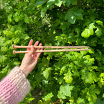 A person holding two wooden knitting needles in front of a bush, ready to start a new knitting project.