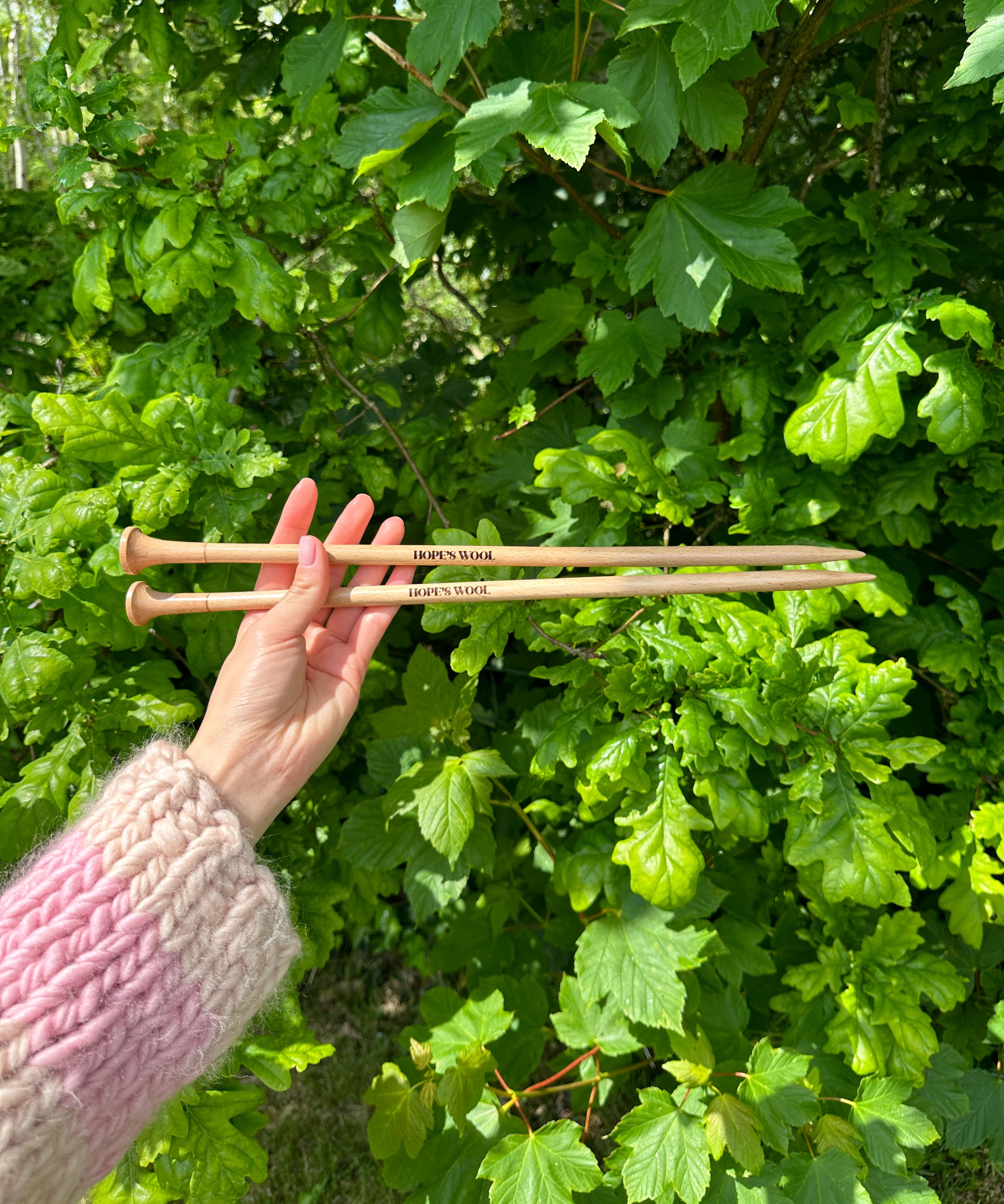 A person holding two wooden knitting needles in front of a bush, ready to start a new knitting project.