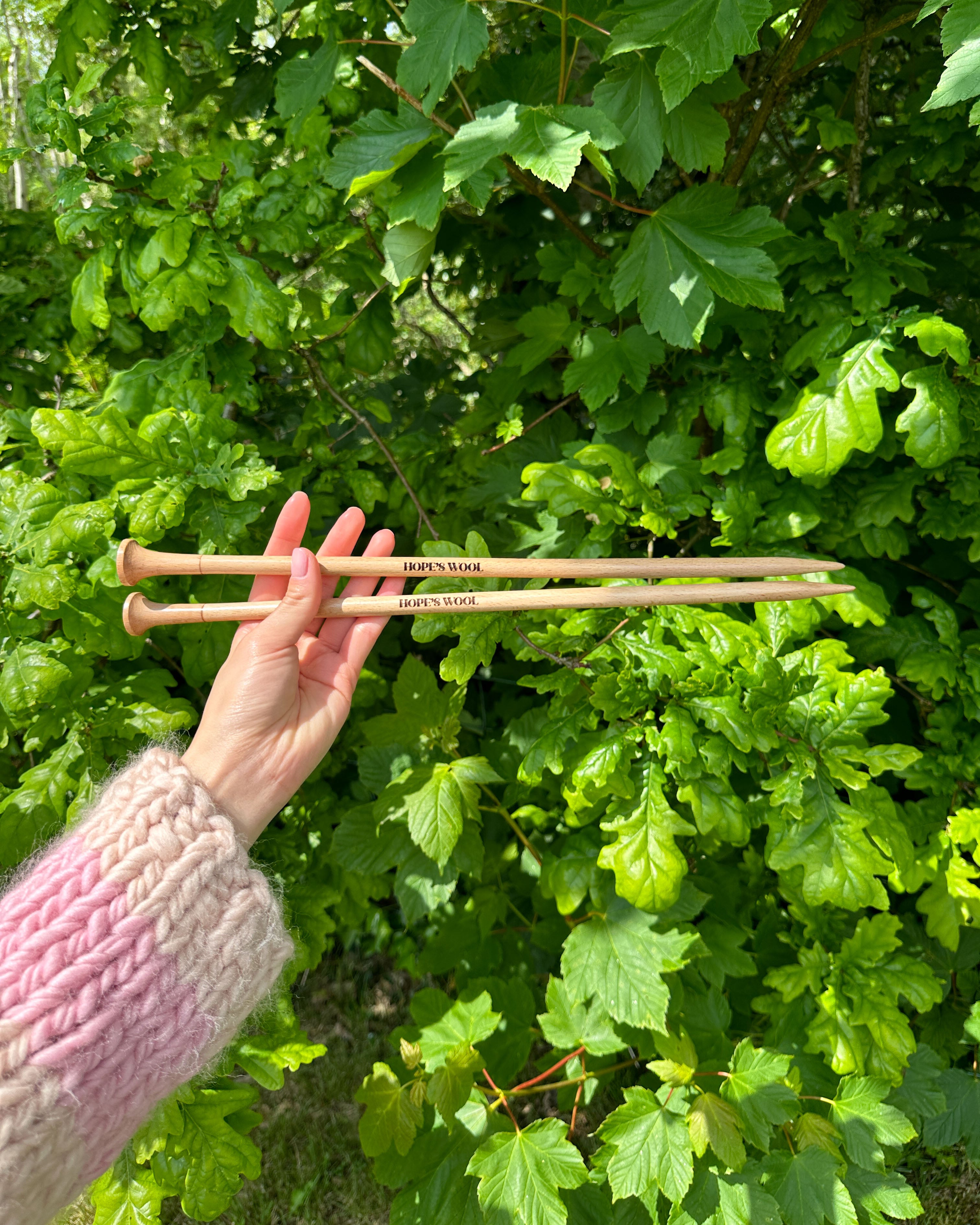 A person holding two wooden knitting needles in front of a bush, ready to start a new knitting project.