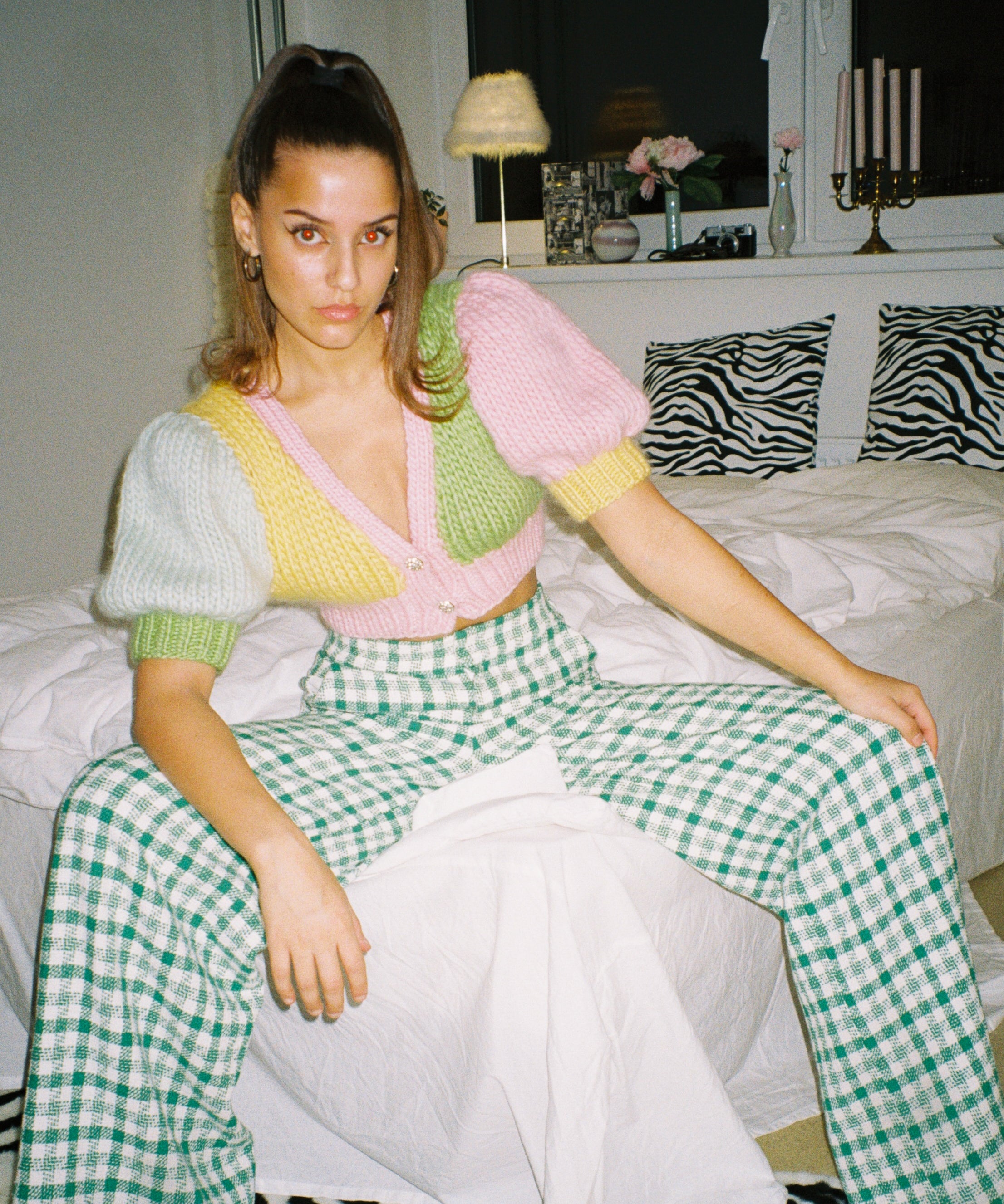 A woman sitting on a bed wearing a colourful knitted top, looking relaxed and cozy.