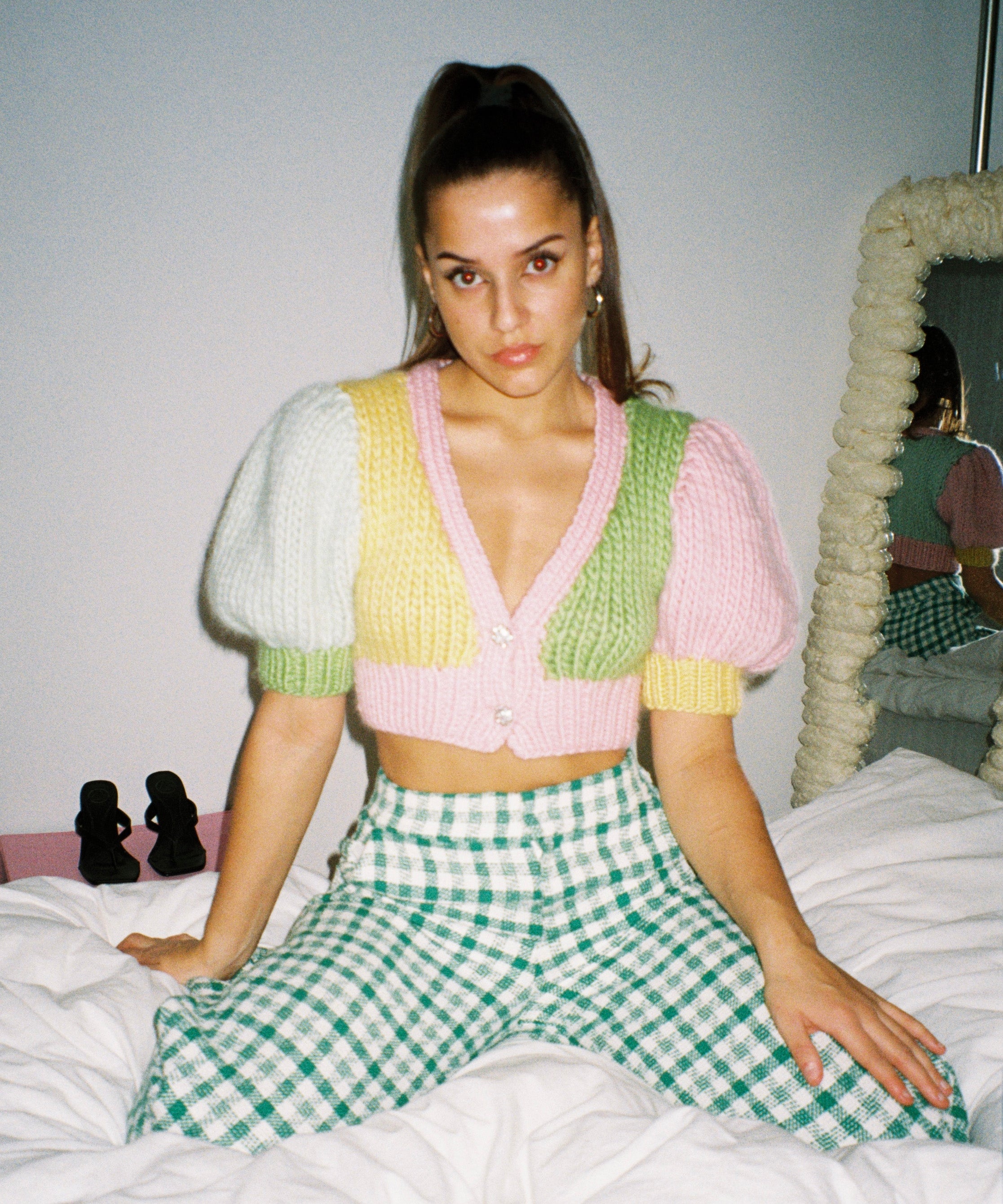 A woman sitting on a bed wearing a colourful knitted top, looking relaxed and cozy.